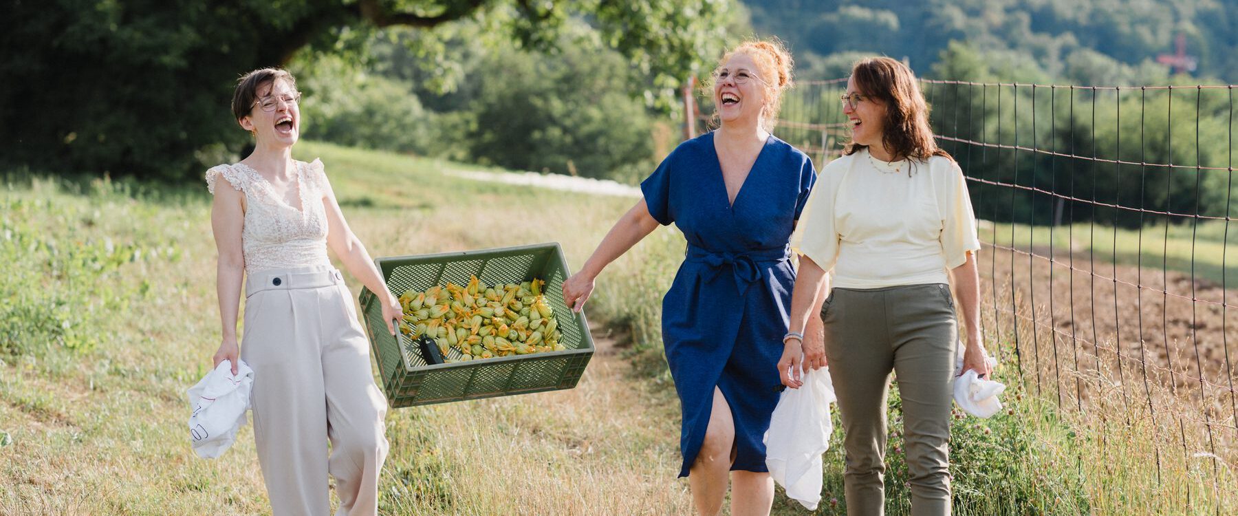 Jacqueline, Christel und Mirjam von Colli Natur Friseure laufen lachend mit einer Kiste Zucchiniblüten über einen Feldweg